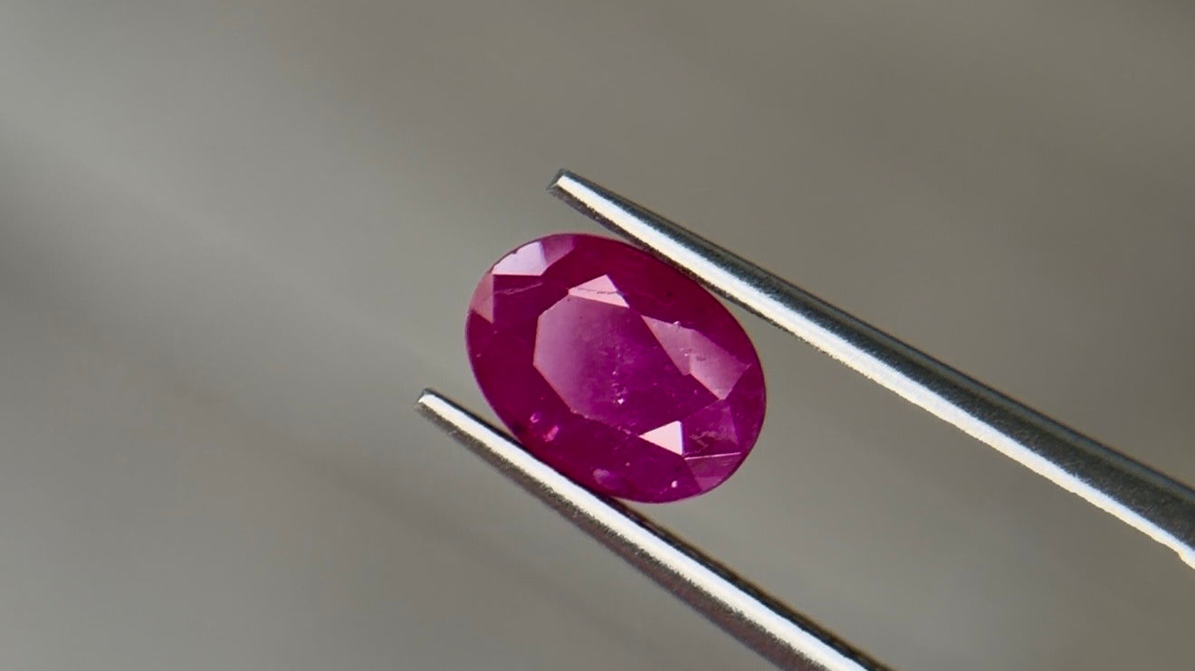 Oval mixed-cut heated Burmese ruby in a medium-dark purplish red with strong saturation, held face-up in steel tweezers at Sapphire Row - the 15th and 40th anniversary gemstone