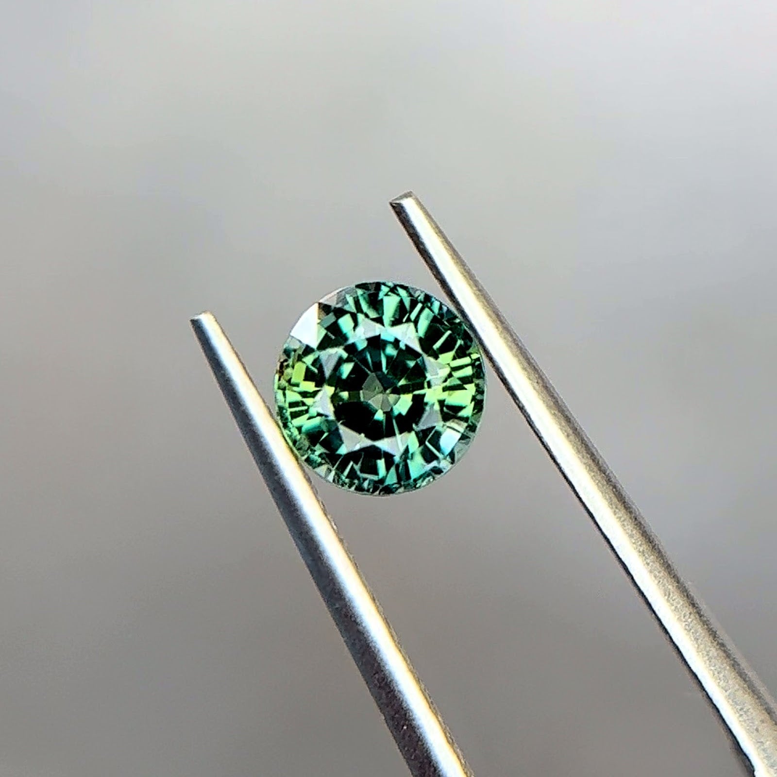 Round brilliant-cut unheated Tanzanian sapphire in a medium bluish green teal, held in stainless steel tweezers at Sapphire Row, showing yellowish green and blue-green color zoning across the crown — the September birthstone beyond classic blue