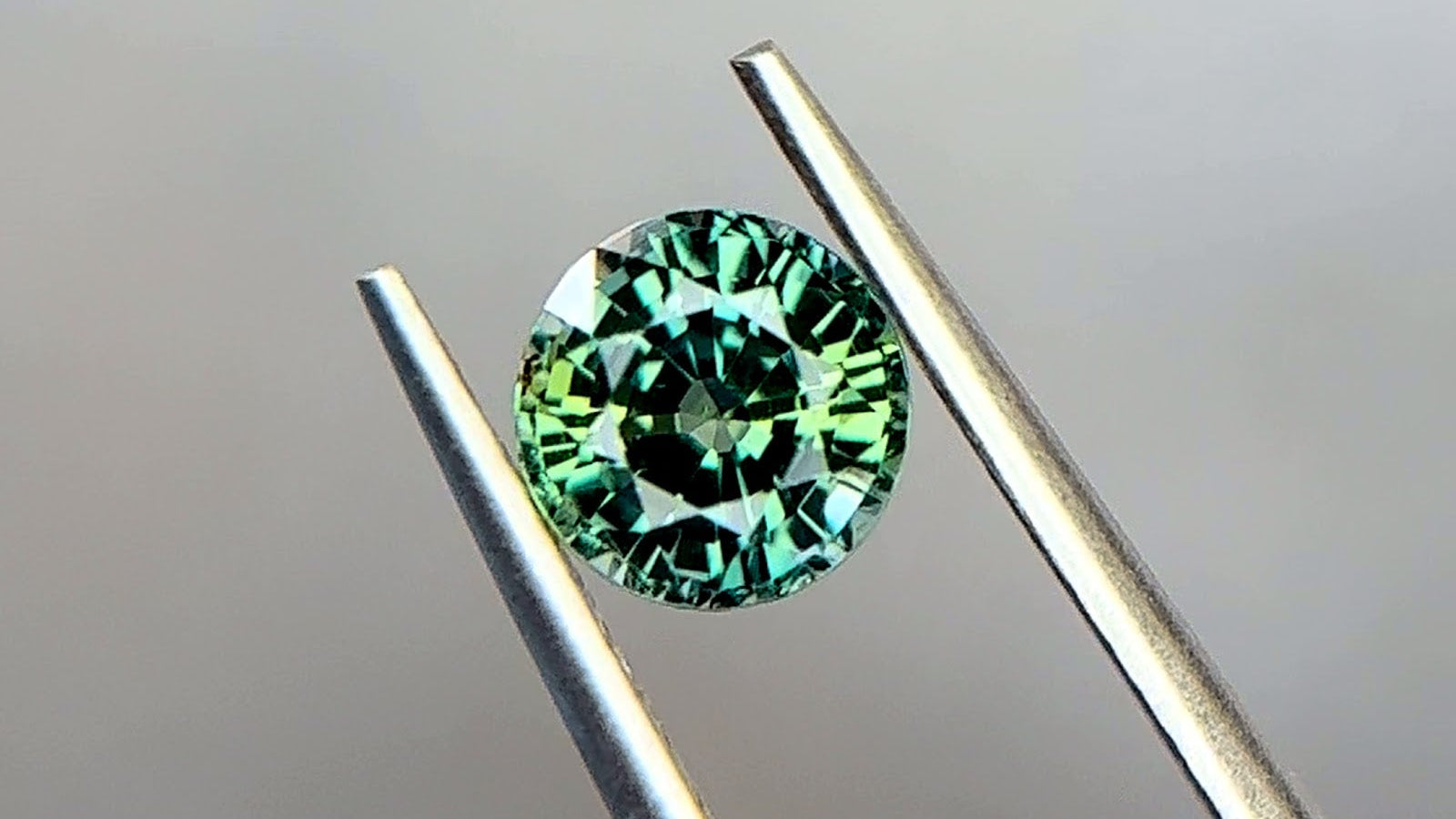 Round brilliant-cut medium bluish green teal sapphire from Tanzania in stainless steel tweezers at Sapphire Row, showing yellowish green and blue-green color zoning, a stone whose gem lab report records species, treatment and origin