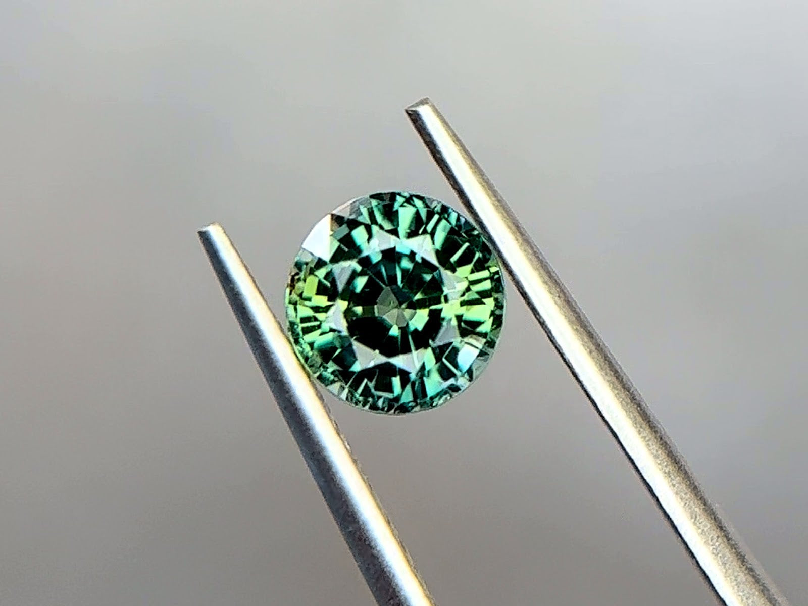 Unheated round brilliant-cut Tanzanian parti sapphire held in stainless steel tweezers at Sapphire Row, showing a medium bluish green teal body with yellowish green and blue-green color zoning across the crown