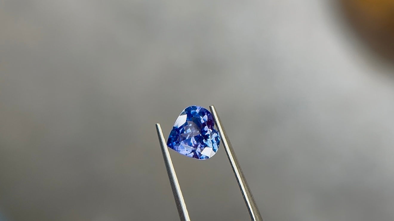 Heart-shaped brilliant-cut tanzanite in a medium to medium-dark violetish blue, held in polished stainless steel tweezers at Sapphire Row, showing violet flashes across the crown facets