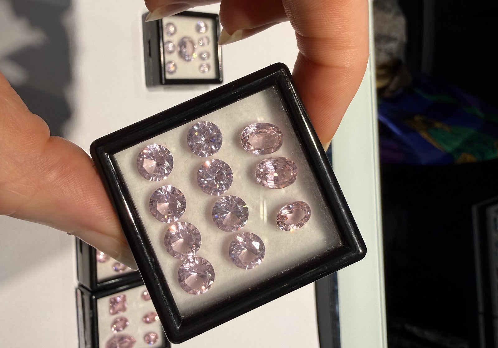 Hand holding a gem box of matched pastel pink oval and round brilliant-cut gemstones at Sapphire Row, an example of the natural colored gemstones discussed in this lab-grown vs natural guide