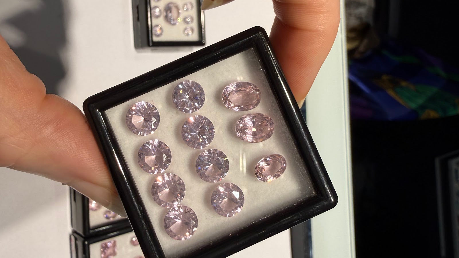 Hand holding a gem box of matched pastel pink oval and round brilliant-cut gemstones at Sapphire Row, an example of the natural colored gemstones discussed in this lab-grown vs natural guide
