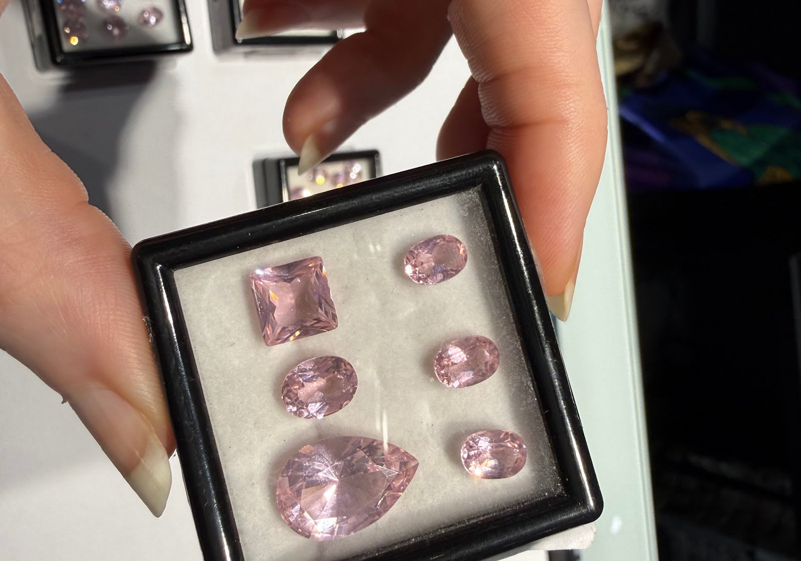 A hand holding an open black gem box of loose pastel pink faceted gemstones, including a square step cut, a large pear and four ovals, with trays of loose colored gemstones behind at Sapphire Row