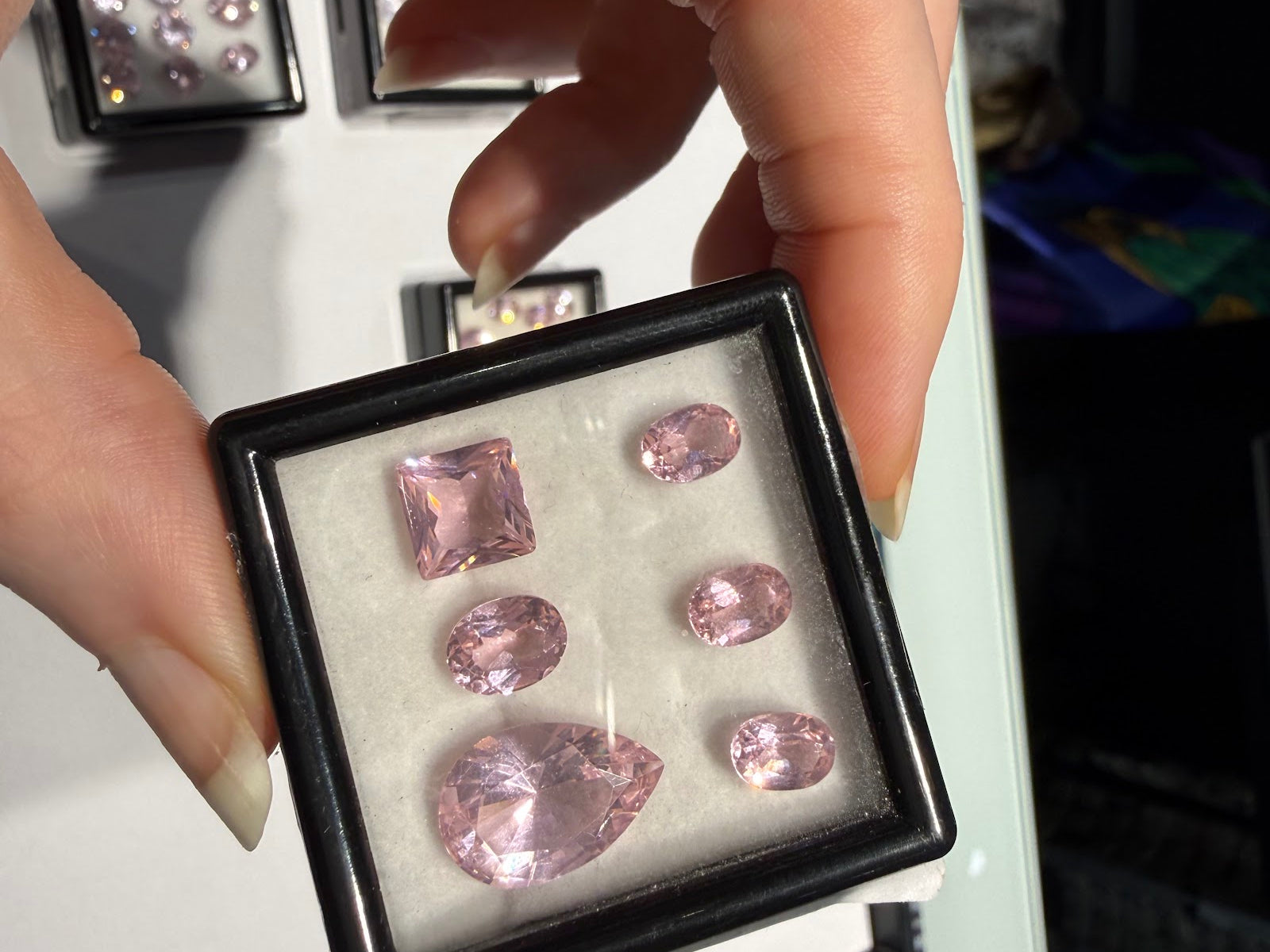 A hand holding an open black gem box of loose pastel pink faceted gemstones, including a square step cut, a large pear and four ovals, with trays of loose colored gemstones behind at Sapphire Row