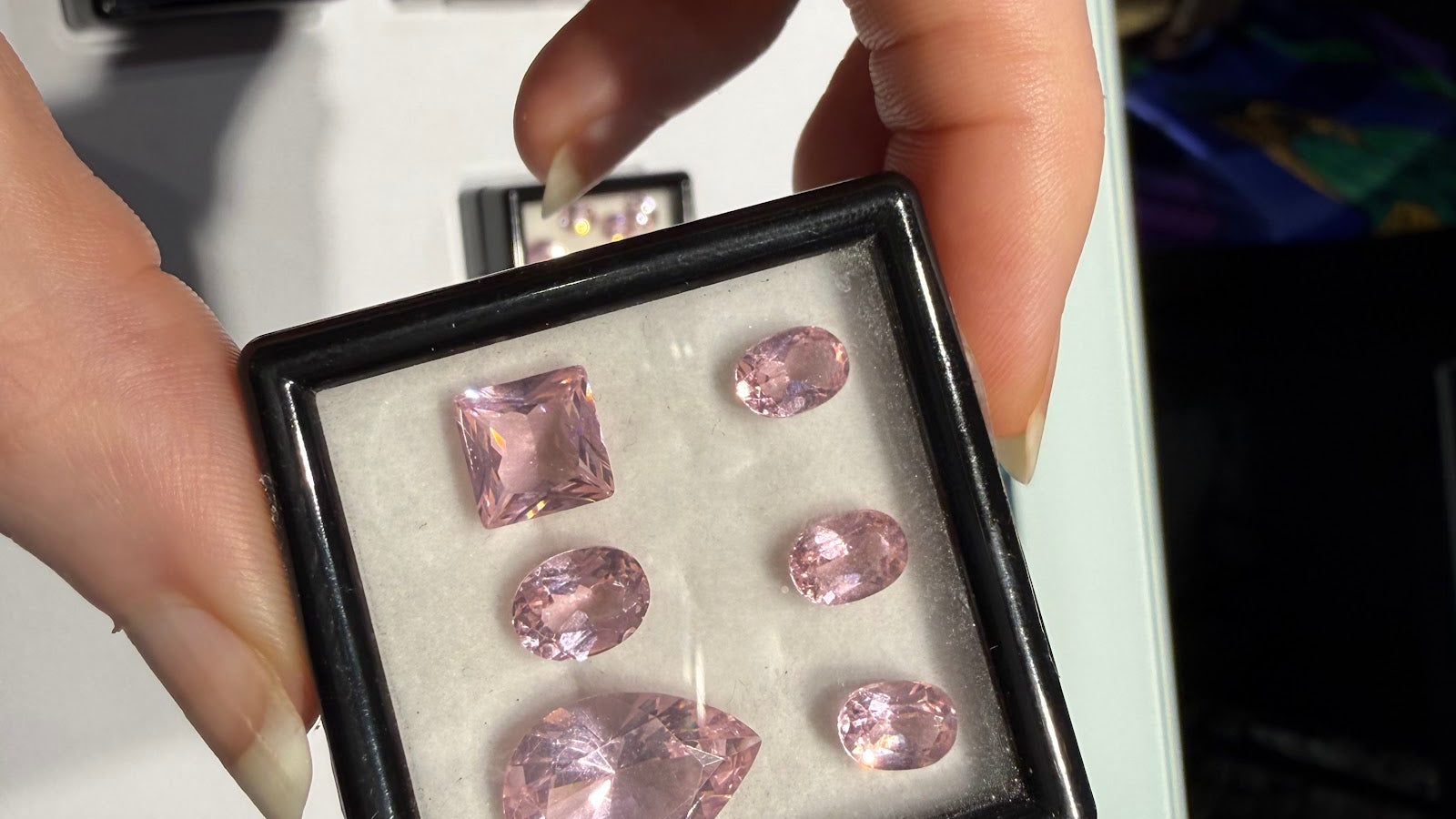 A hand holding an open black gem box of loose pastel pink faceted gemstones, including a square step cut, a large pear and four ovals, with trays of loose colored gemstones behind at Sapphire Row