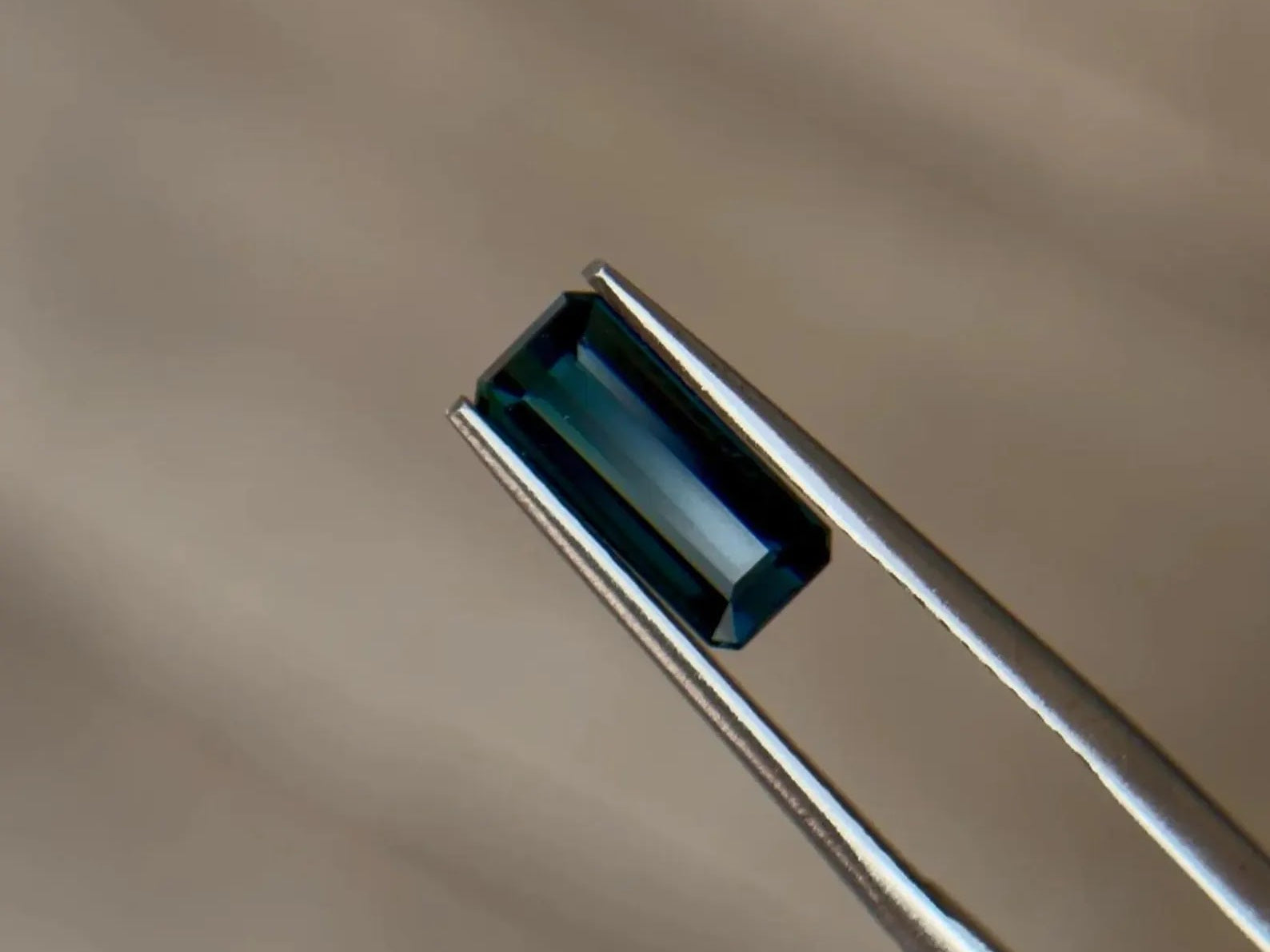 Emerald-cut teal blue sapphire held in jeweler's tweezers, showcasing the durability of corundum gemstones