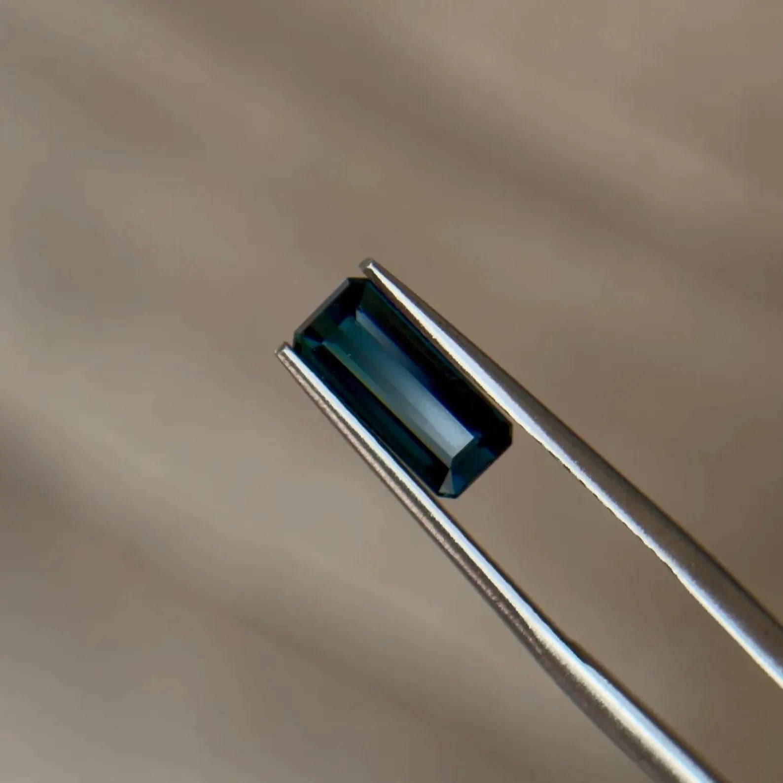 Emerald-cut teal blue sapphire held in jeweler's tweezers, showcasing the durability of corundum gemstones