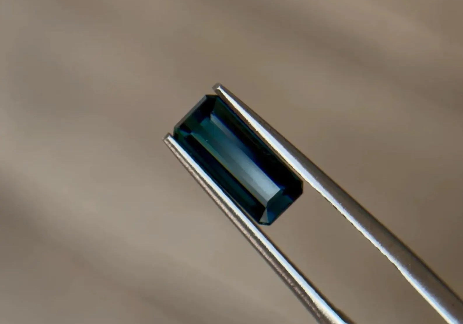 Elongated emerald-cut teal blue-green natural sapphire held in stainless steel tweezers against a soft neutral background at Sapphire Row