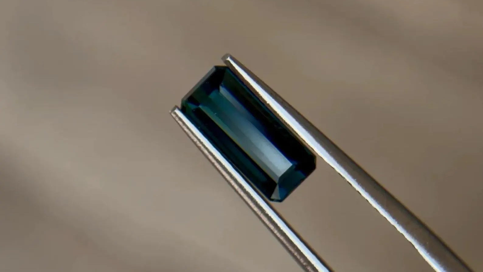 Emerald-cut teal-blue natural sapphire held in jeweler's tweezers at Sapphire Row, a finely cut stone destined for a hallmarked precious-metal setting