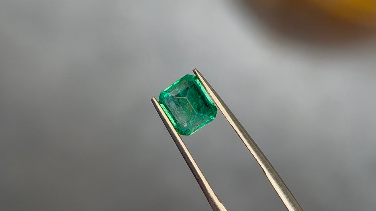 Emerald-cut vivid slightly bluish green Zambian emerald held in warm gold-toned tweezers at Sapphire Row, showing whitish jardin veils and mossy internal texture across the table