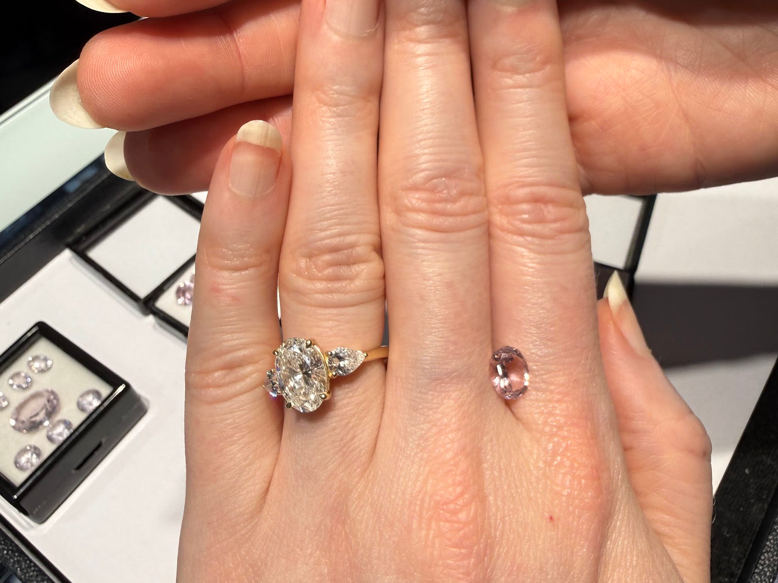 A hand wearing an oval three-stone engagement ring in yellow gold beside a loose oval pink gemstone, with trays of loose colored gemstones at Sapphire Row