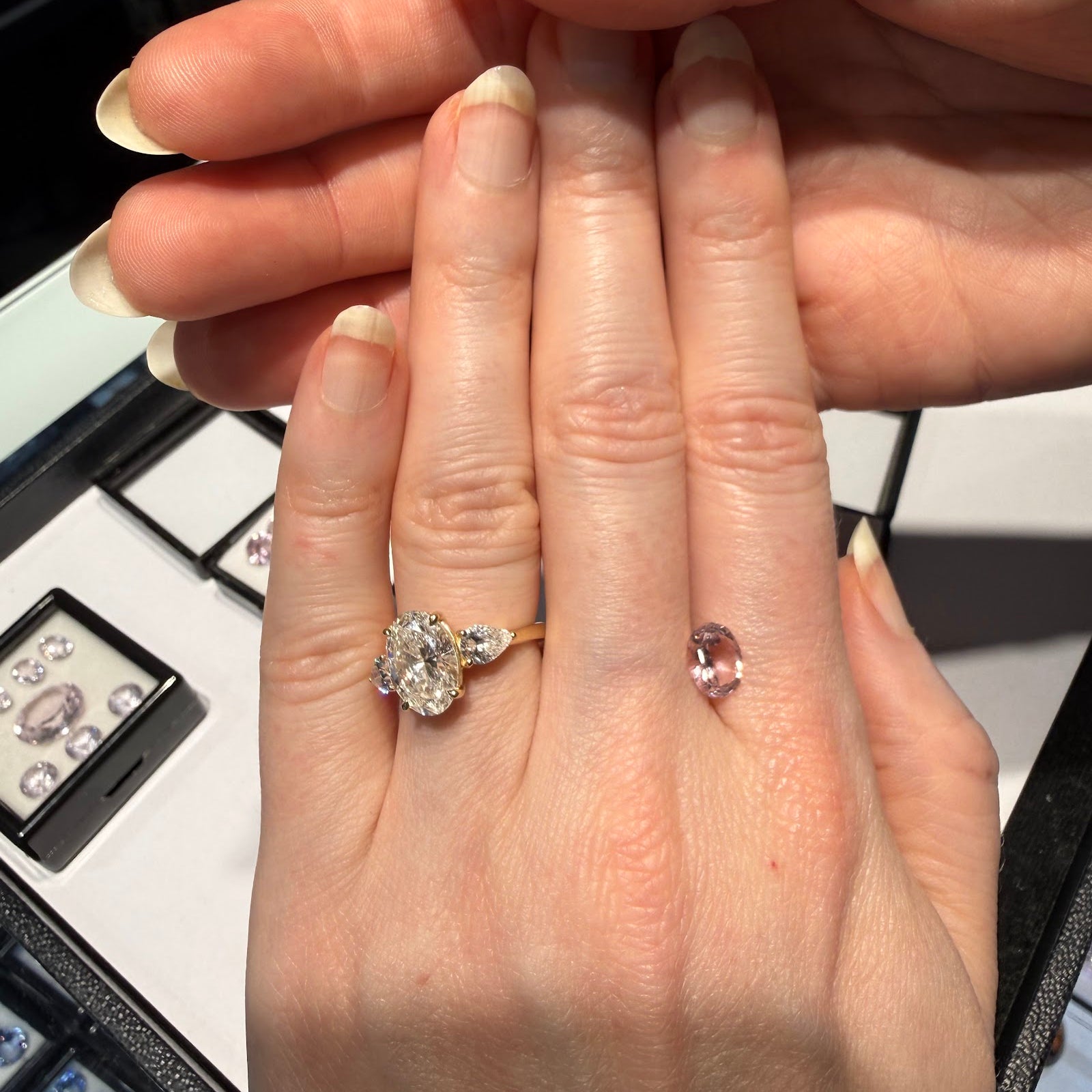 A hand wearing an oval three-stone engagement ring in yellow gold beside a loose oval pink gemstone, with trays of loose colored gemstones at Sapphire Row