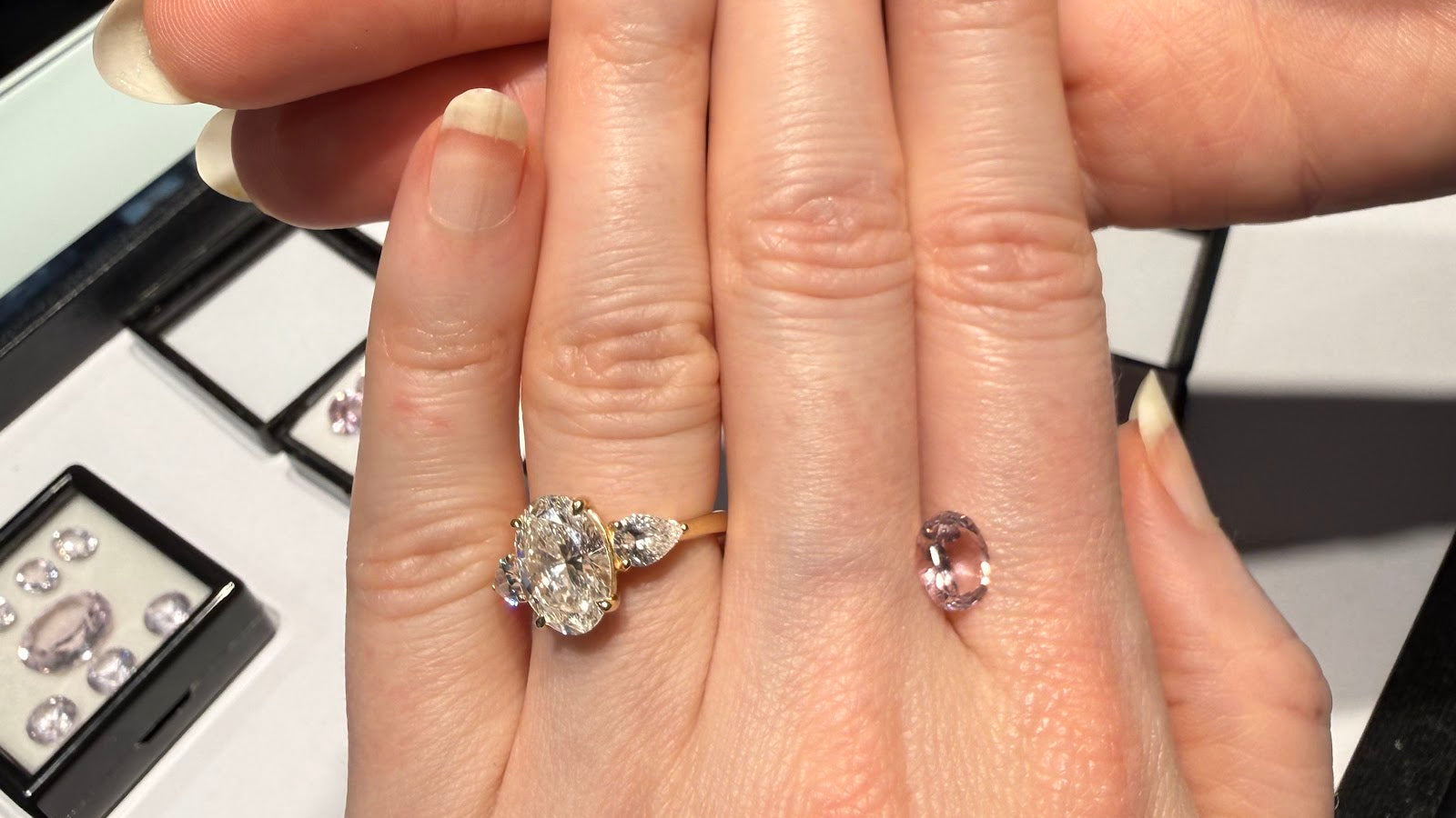 A hand wearing an oval three-stone engagement ring in yellow gold beside a loose oval pink gemstone, with trays of loose colored gemstones at Sapphire Row