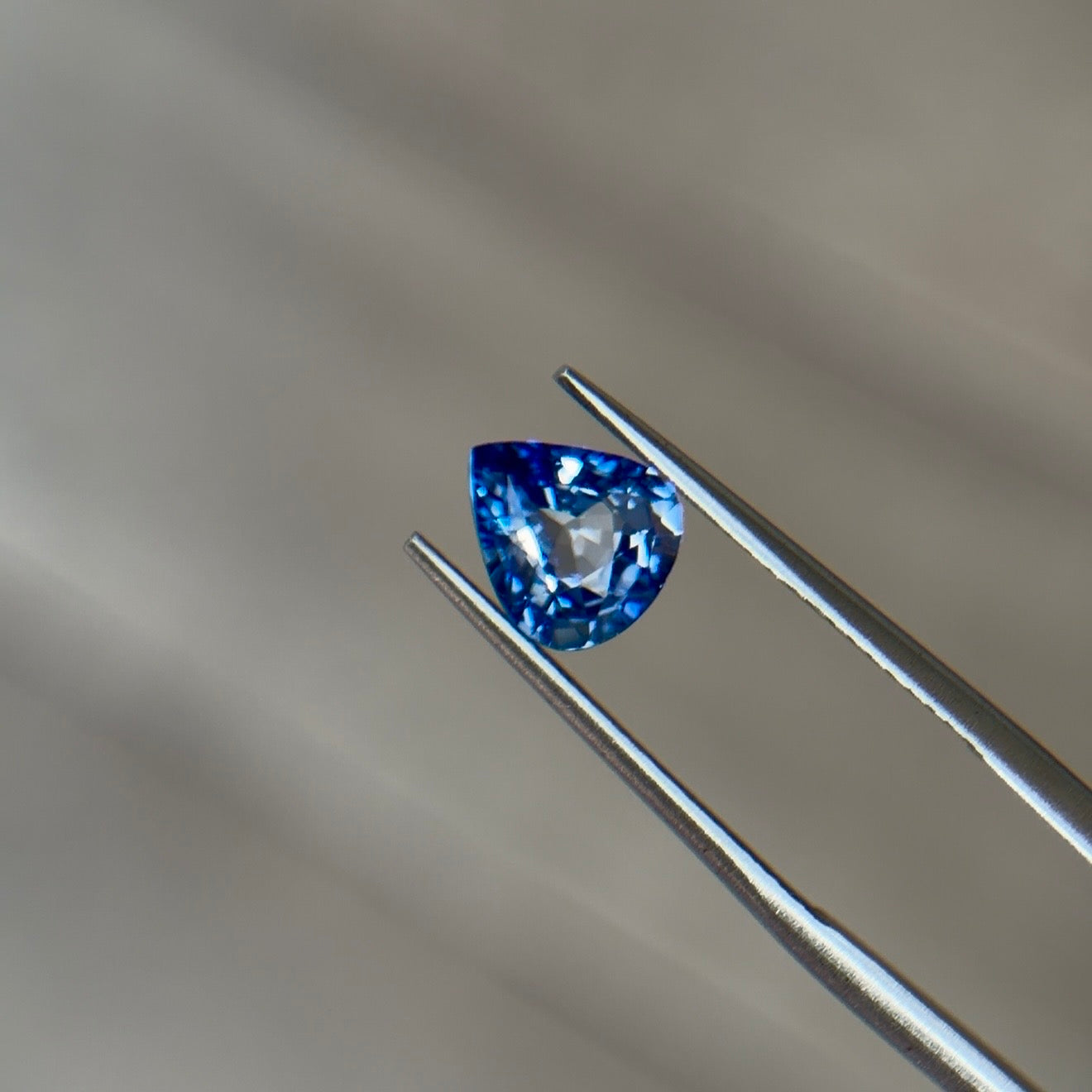 Pear-shaped brilliant-cut sapphire in a medium slightly violetish blue held in steel tweezers at Sapphire Row, a hard, tough corundum ideal for a gemstone engagement ring