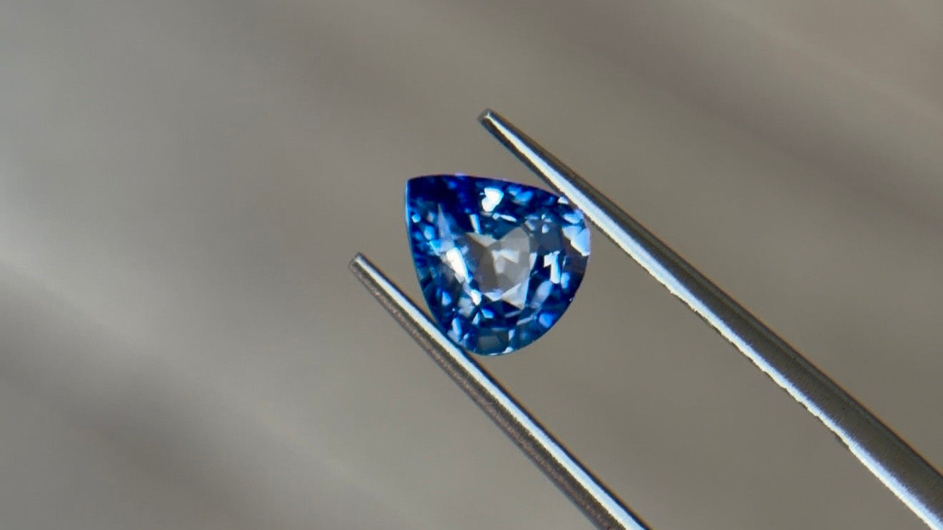 Pear-shaped brilliant-cut sapphire in a medium slightly violetish blue held in steel tweezers at Sapphire Row, a hard, tough corundum ideal for a gemstone engagement ring