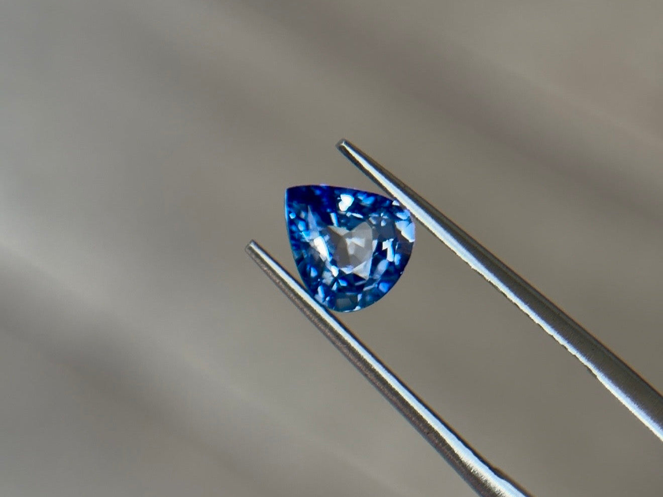 Pear-shaped brilliant-cut sapphire in a medium slightly violetish blue held in steel tweezers at Sapphire Row, a hard, tough corundum ideal for a gemstone engagement ring
