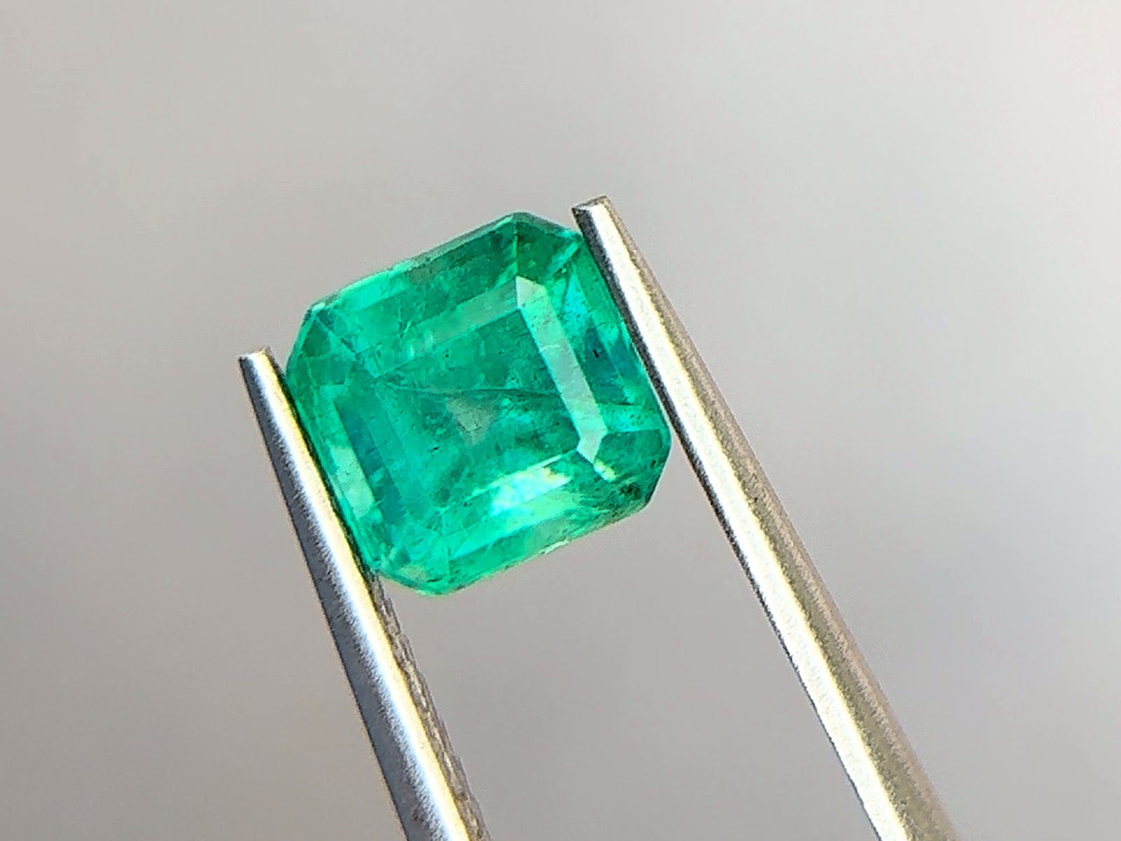 Emerald-cut Zambian emerald in a medium slightly bluish green with strong saturation, held face-up in polished steel tweezers at Sapphire Row, showing wispy jardin veils and healed fissures across the table
