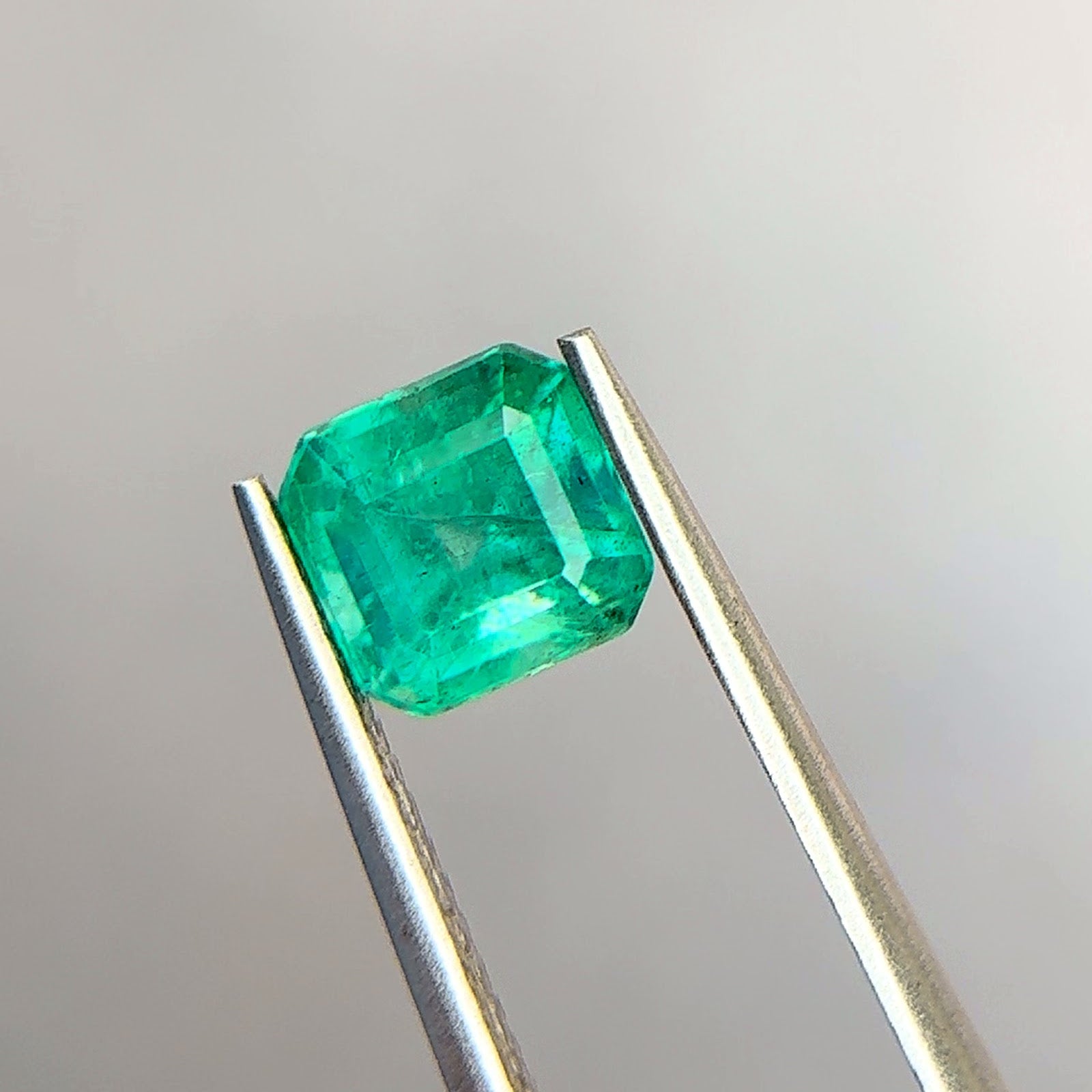 Emerald-cut Zambian emerald in a medium slightly bluish green with strong saturation, held face-up in polished steel tweezers at Sapphire Row, showing wispy jardin veils and healed fissures across the table