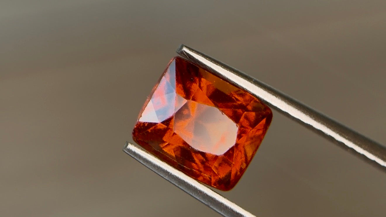 Cushion-cut natural hessonite garnet in a vivid reddish orange with fiery flashes, held tilted in steel tweezers at Sapphire Row - garnet, the January birthstone, beyond classic red