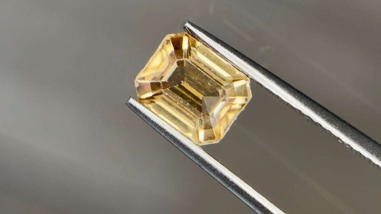 Medium slightly brownish to orangy yellow natural zircon in a 1.97 carat octagon step cut, held in tweezers against a warm gray background