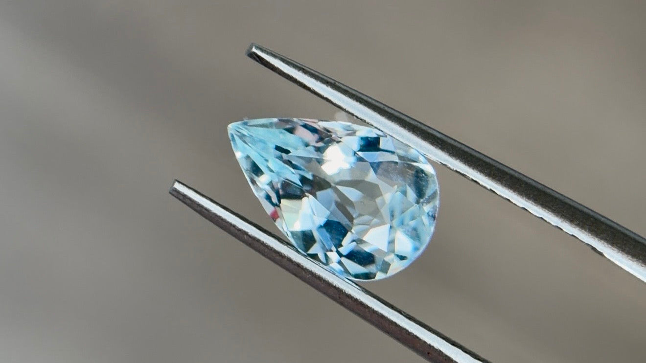 Pear-shaped aquamarine in light to medium slightly greenish blue held in tweezers, the modern March birthstone
