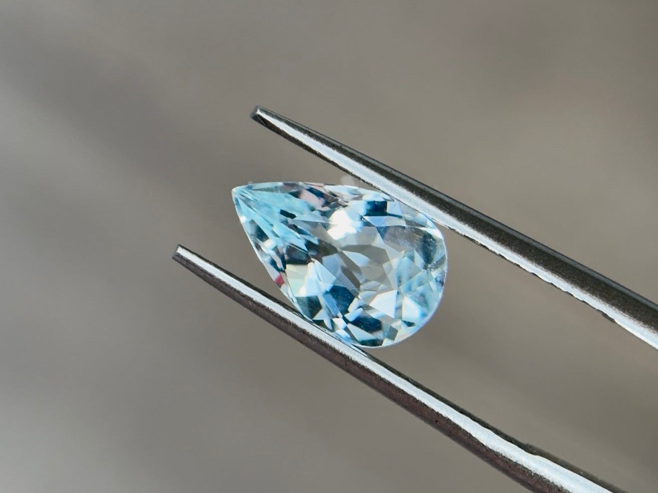 Pear-shaped aquamarine in light to medium slightly greenish blue held in tweezers, the modern March birthstone