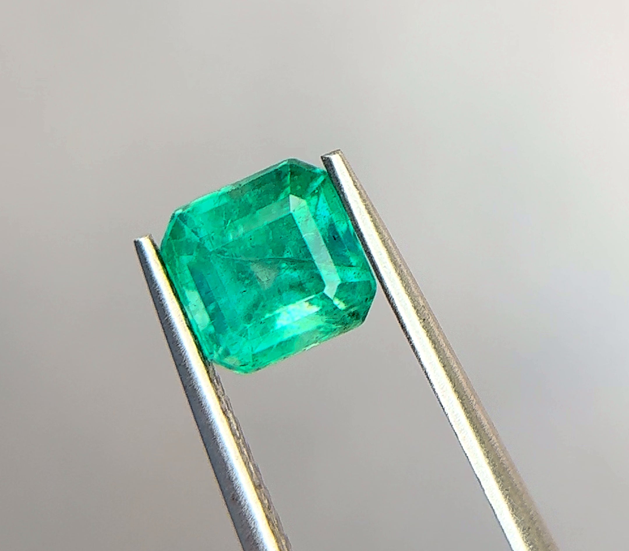 Vivid slightly bluish green emerald-cut emerald held in tweezers against a neutral background, a stone that benefits from a protective bezel or corner prongs