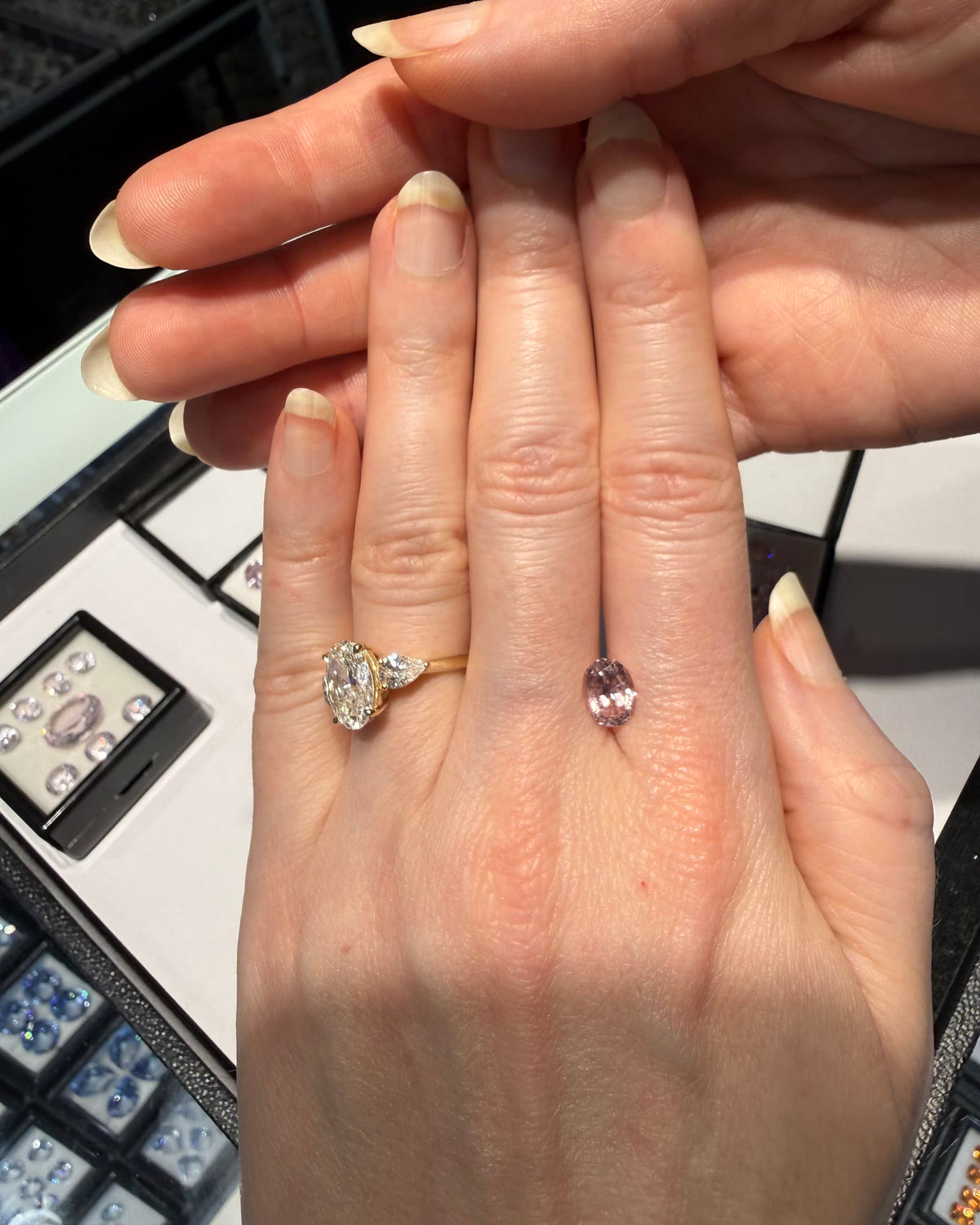 An oval diamond ring with a pear side stone in yellow gold, worn beside a loose pink gemstone