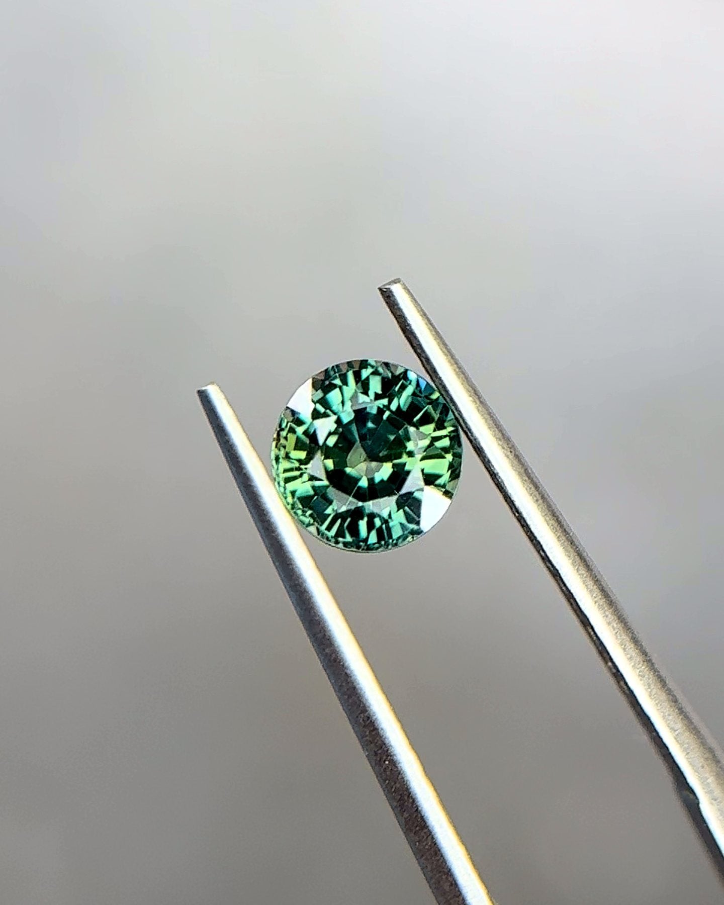 A parti sapphire in bluish green and yellowish green, held in tweezers for a closer look